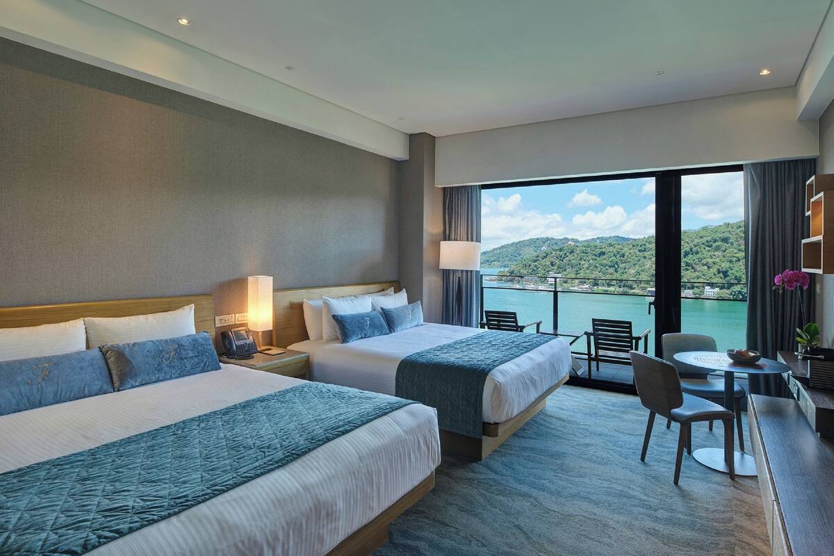 Habitación de hotel con dos camas, un escritorio con silla, y vista a un cuerpo de agua y colinas.