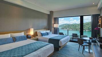 A hotel room with two beds, a desk with a chair, and a view of a body of water and hills.