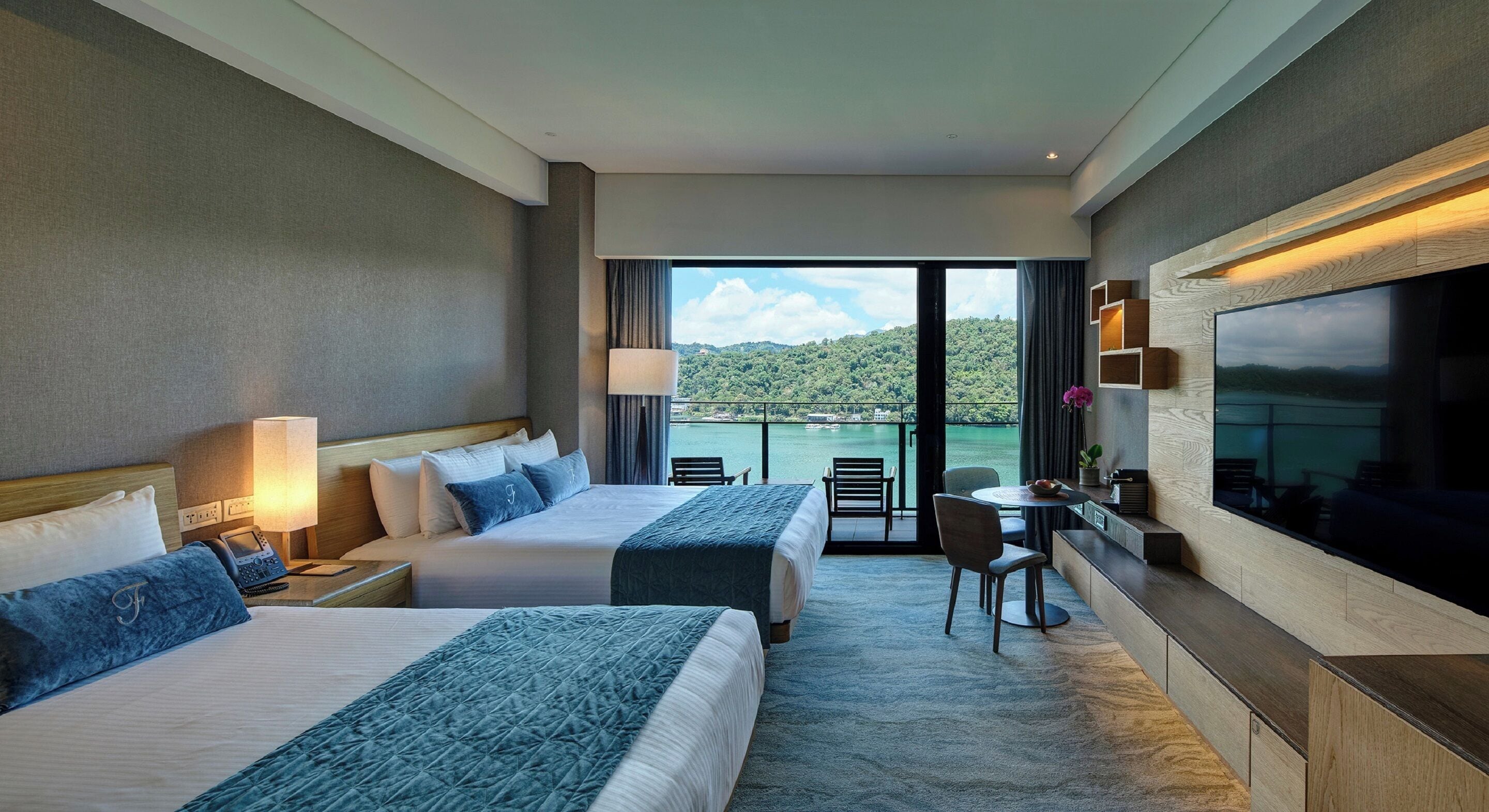 A hotel room with two beds, a desk, chairs, a TV, and a balcony with a view of the water.