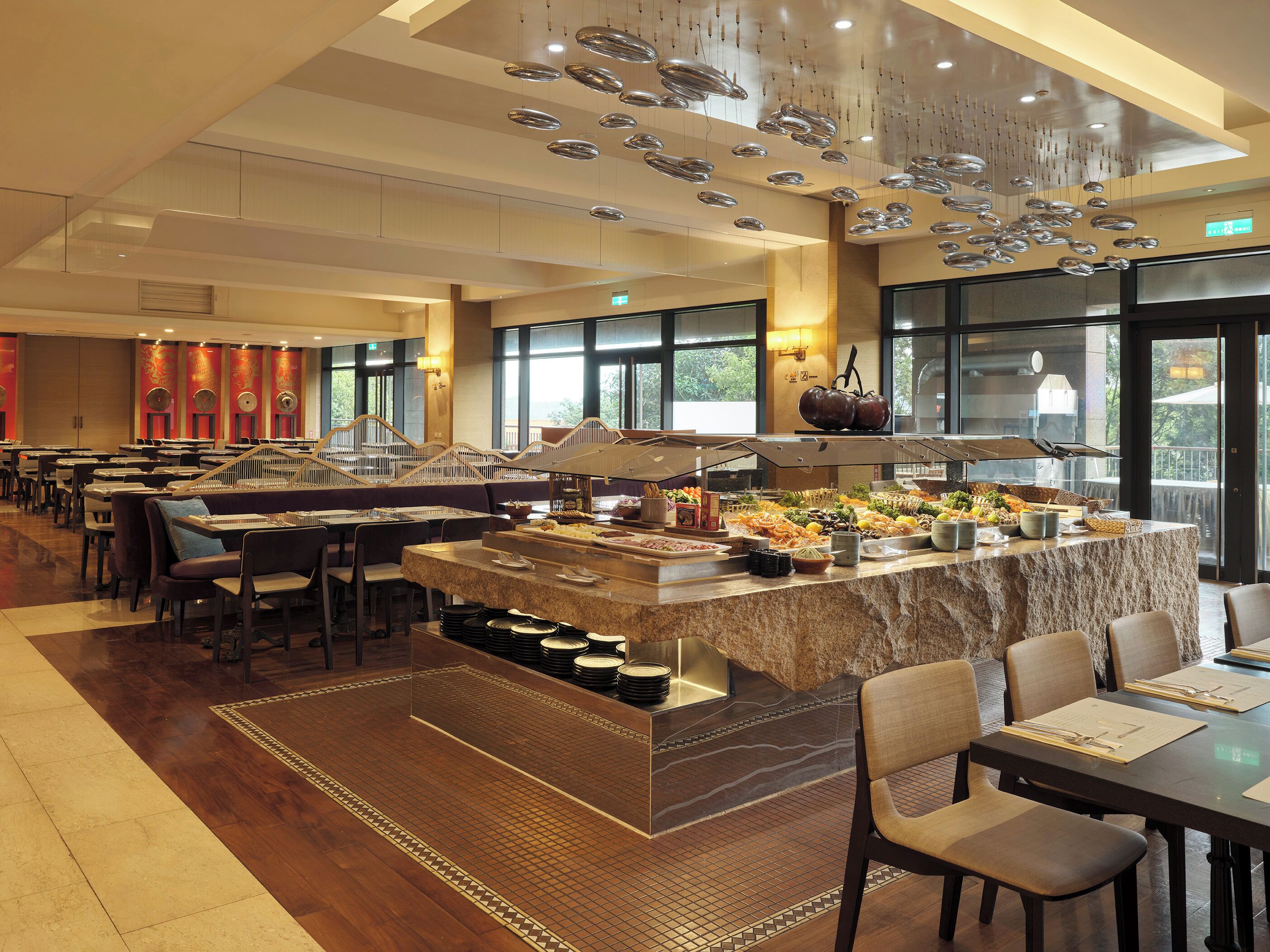 Un comedor amplio con varias mesas y sillas, una barra de buffet con diversos alimentos, y un ventanal que ofrece vistas al exterior.