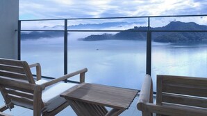 Varanda com cadeiras e mesa de madeira, com vista para um lago calmo e montanhas ao longe.