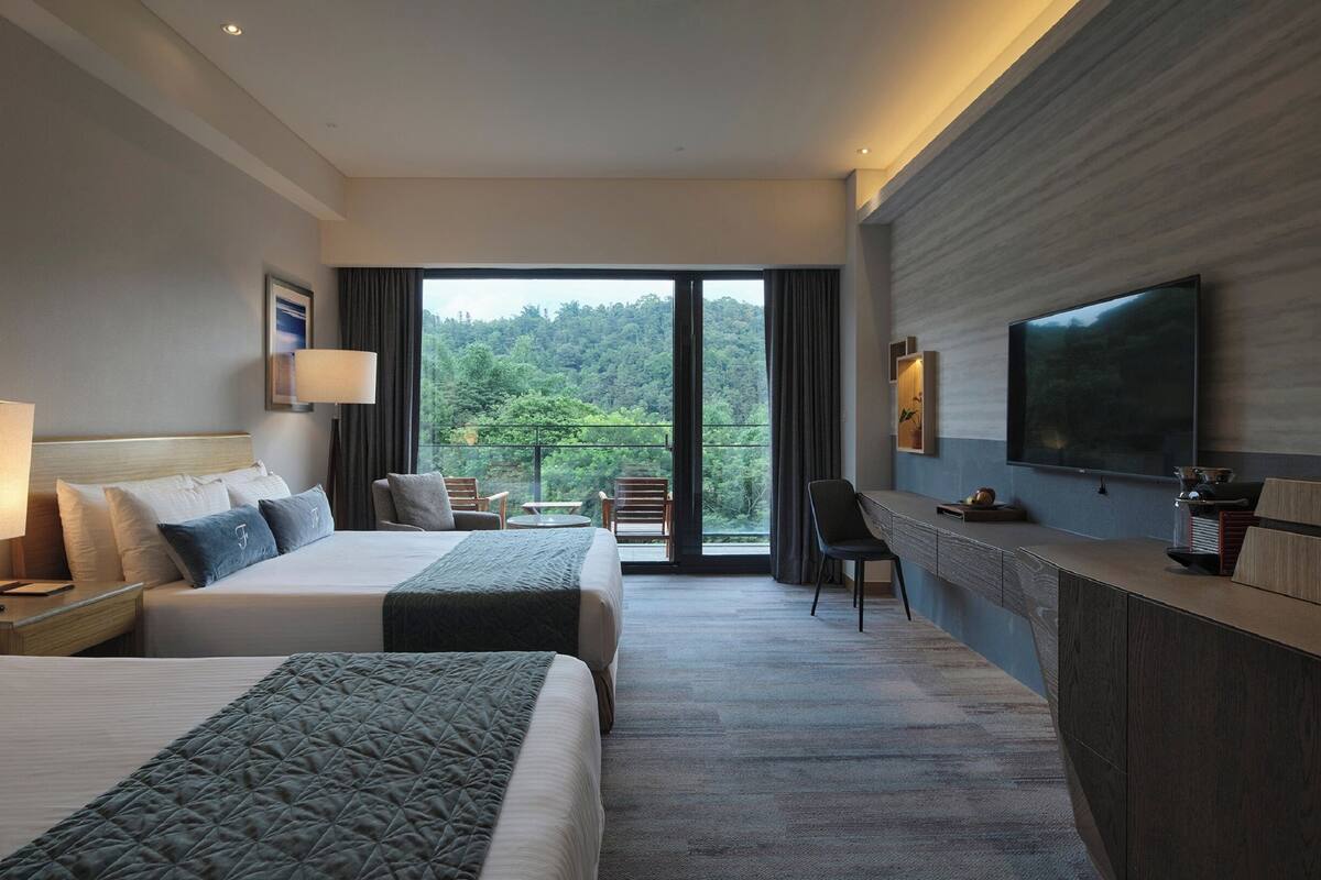 Habitación de hotel con dos camas, un escritorio, televisión y balcón con vistas al bosque.