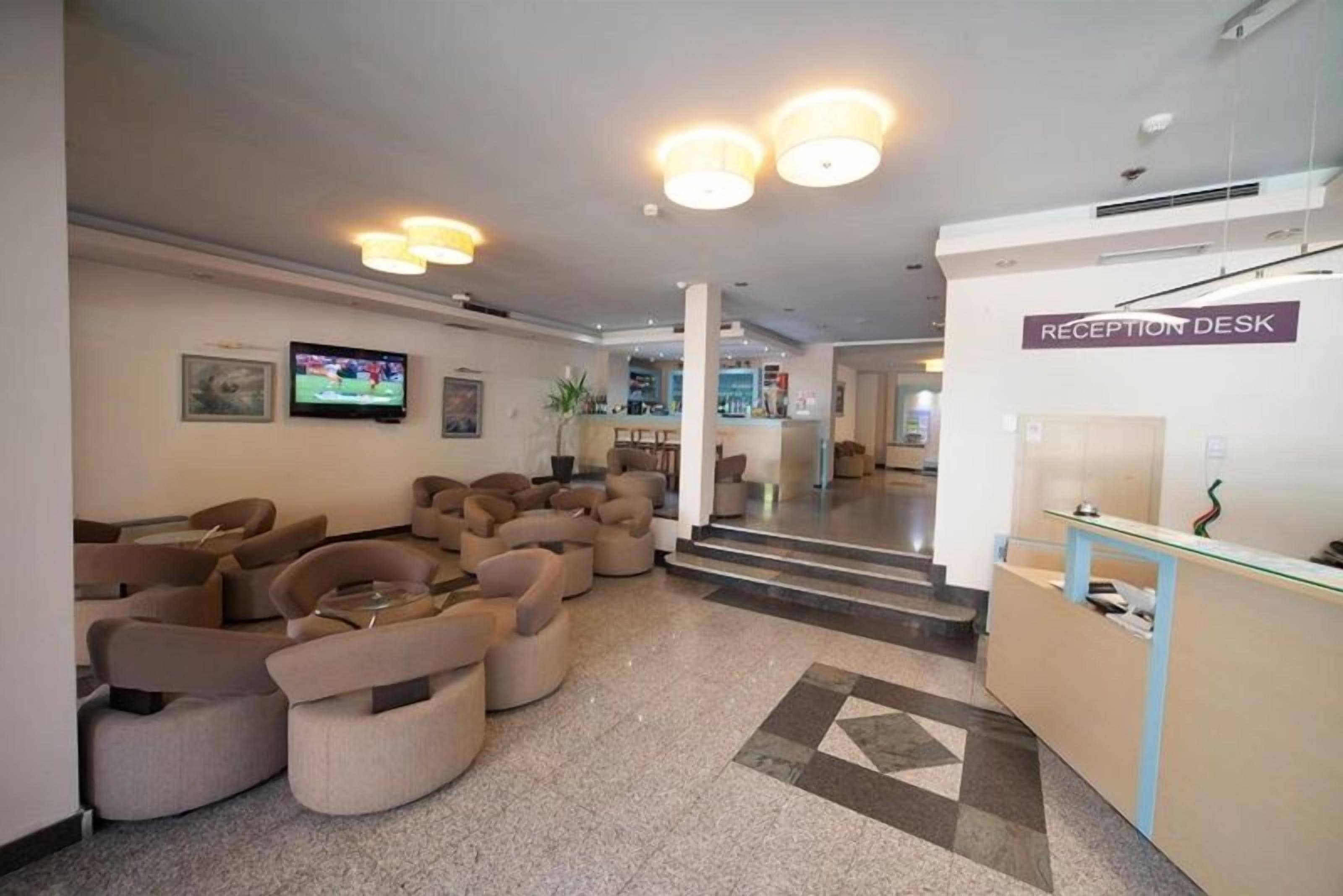 Un vestíbulo de hotel con mostrador de recepción, sofás, televisor y una escalera que lleva a otras áreas.