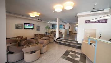 Un vestíbulo de hotel con mostrador de recepción, sofás, televisor y una escalera que lleva a otras áreas.