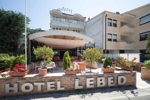 Entrada de un hotel con un letrero que dice 'HOTEL LEBED', mesas al aire libre y plantas en macetas.