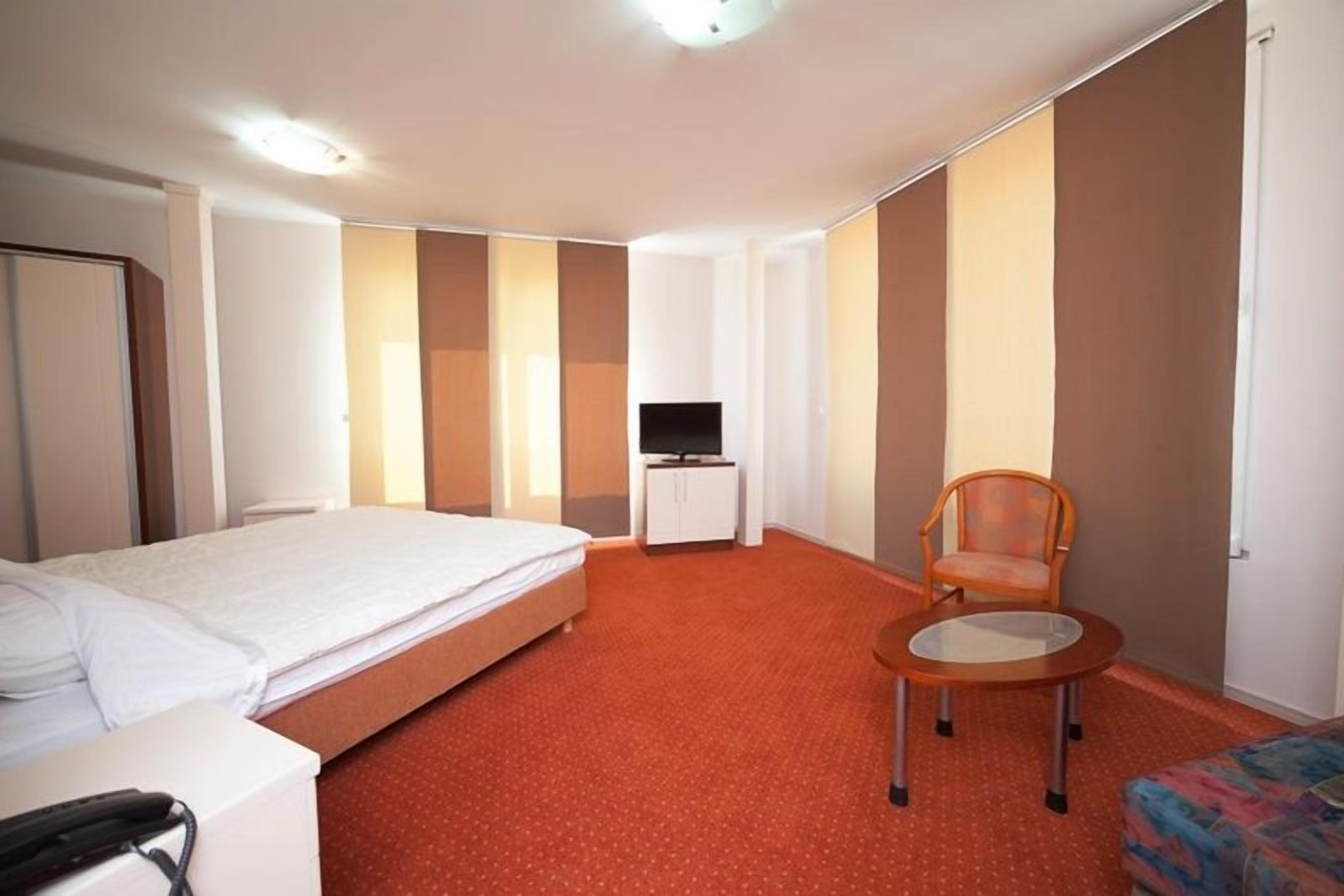 A hotel room with a bed, a small table, a chair, a television, and a wardrobe.