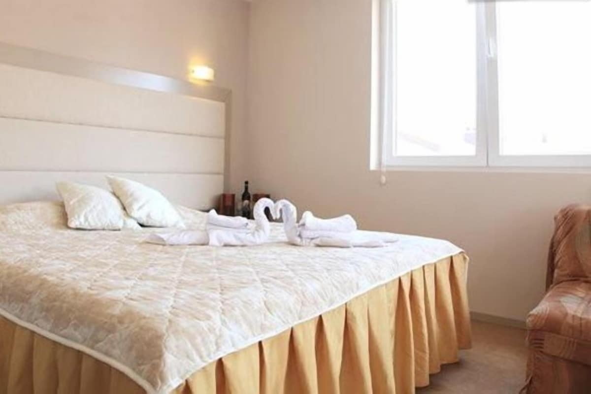 A neatly made bed with a beige bedspread and pillows, a bedside table with a bottle and glasses, and a window providing natural light.