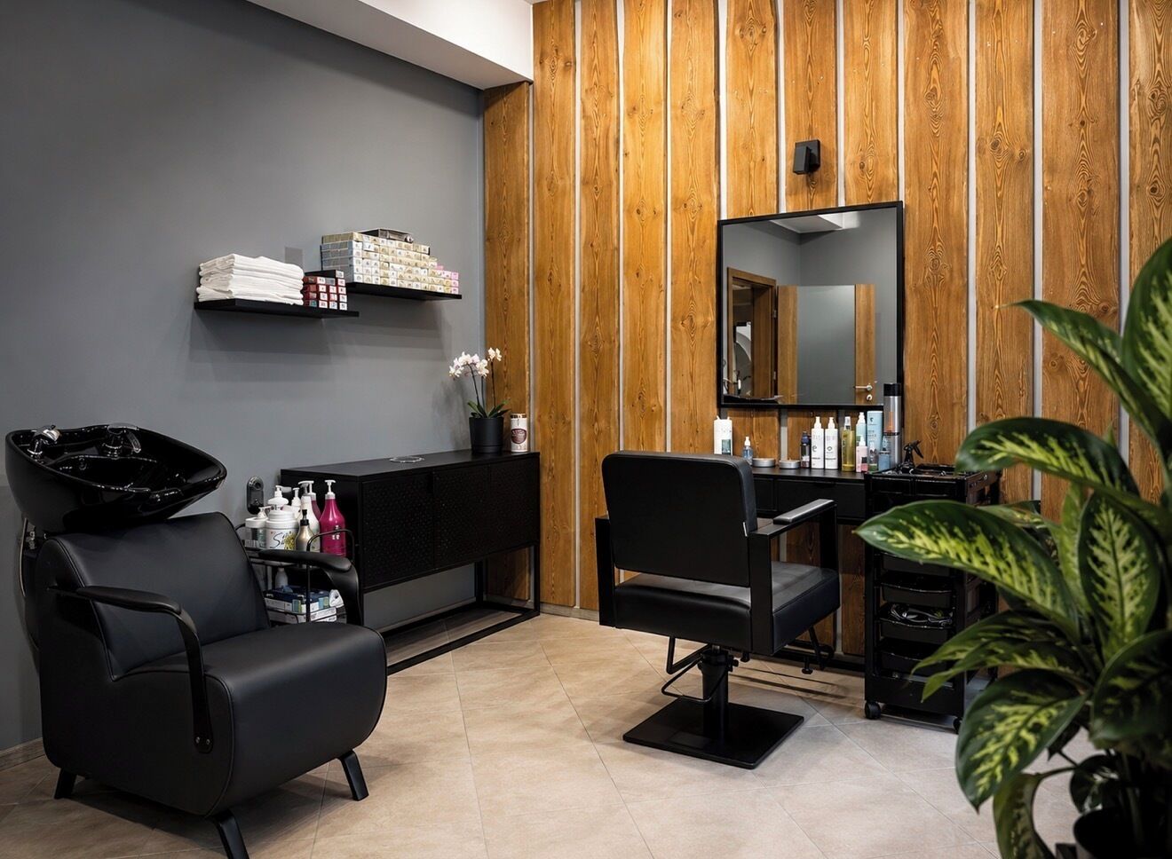 Un salón moderno con dos sillas de peluquería negras, un panel de pared de madera, un espejo y una estantería con productos para el cabello.
