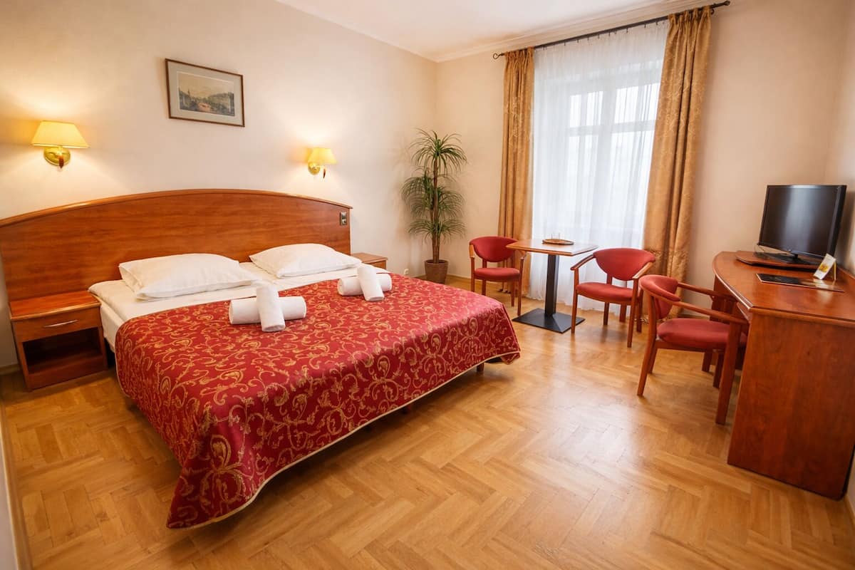 A hotel room with a wooden bed, a desk with a TV, two chairs, a small table, and a potted plant.