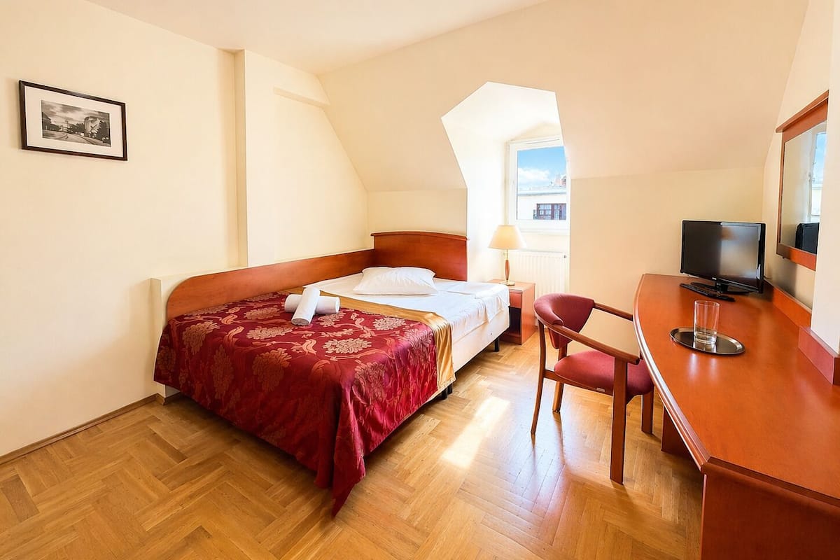 A bedroom with a bed, a desk, a chair, a television, and a window.