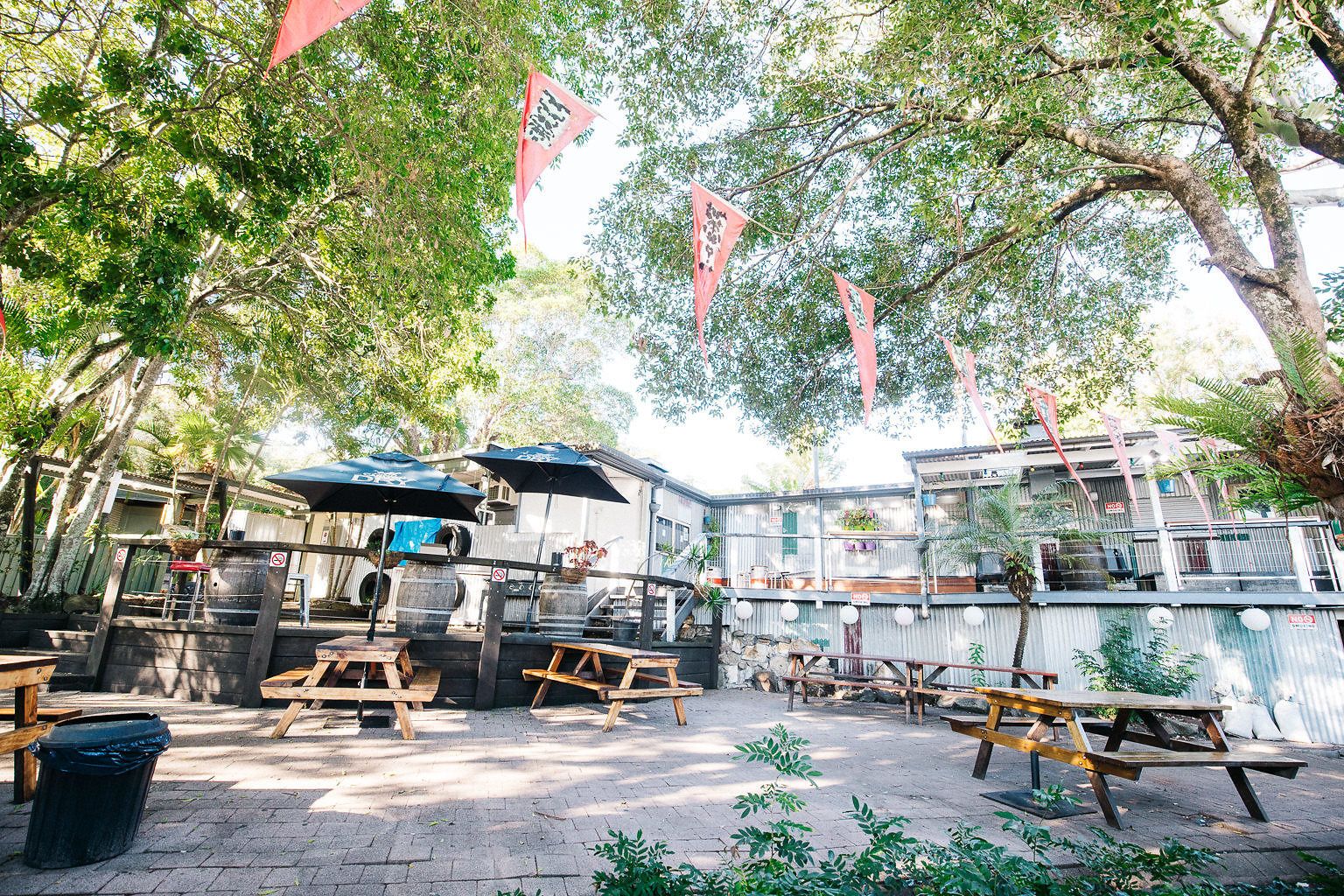 Un espace extérieur aménagé avec des tables et des bancs en bois, entouré d’arbres et bordé d’un bâtiment en métal ondulé.
