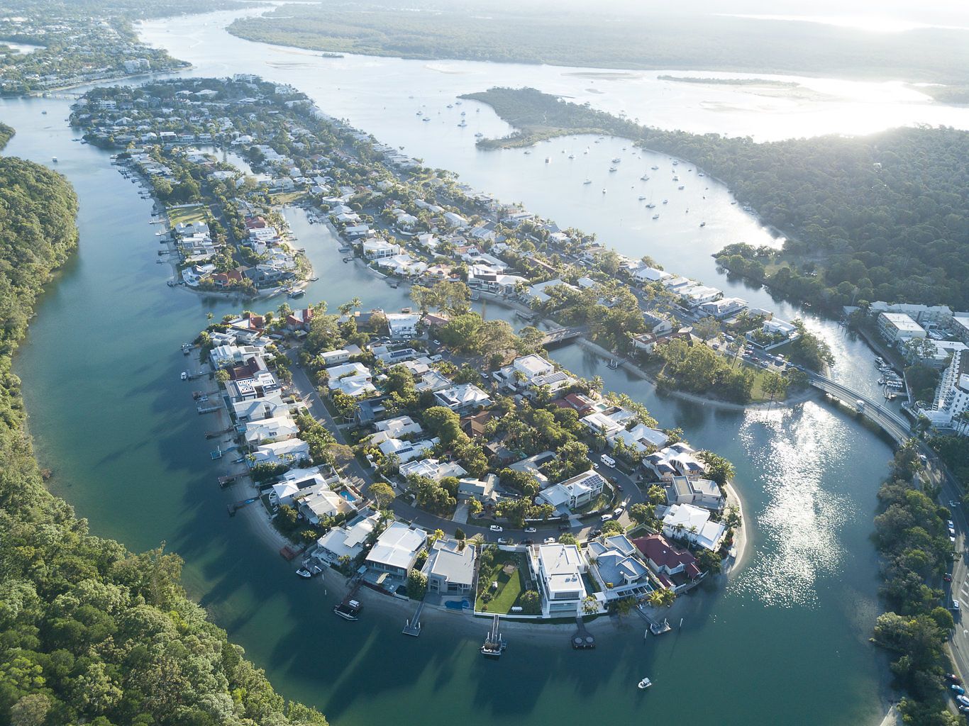 An aerial view of a coastal residential area with houses along the shoreline, a marina with boats, and a bridge connecting the area.