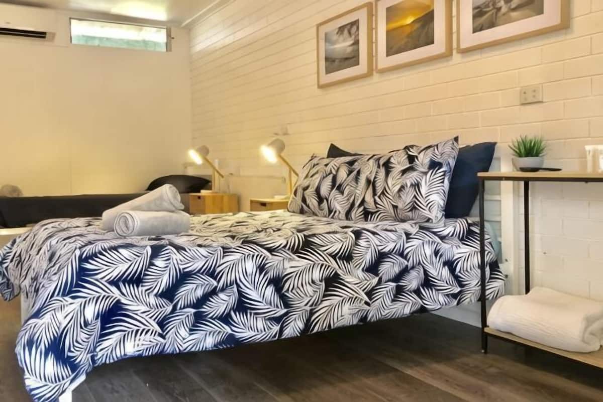 A neatly arranged bedroom with a bed, bedside tables, a small plant, and a wall-mounted shelf.