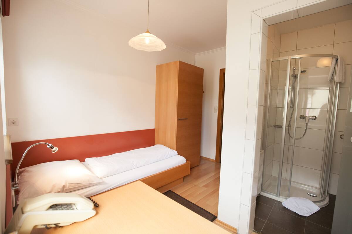 A hotel room with a bed, bedside telephone, wooden wardrobe, and a glass-enclosed shower.