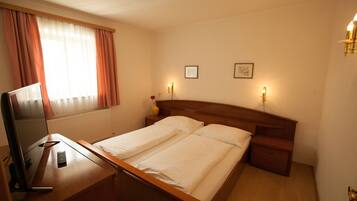 A double bed with white linens, a wooden headboard, bedside tables with lamps, a desk with a computer, and a window with curtains.