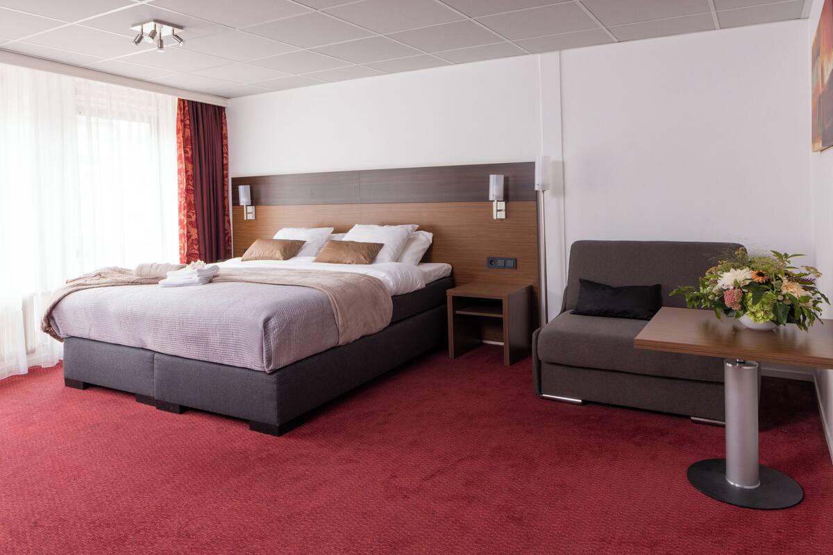 A hotel room with a bed, a sofa, a table with a vase of flowers, and a nightstand.