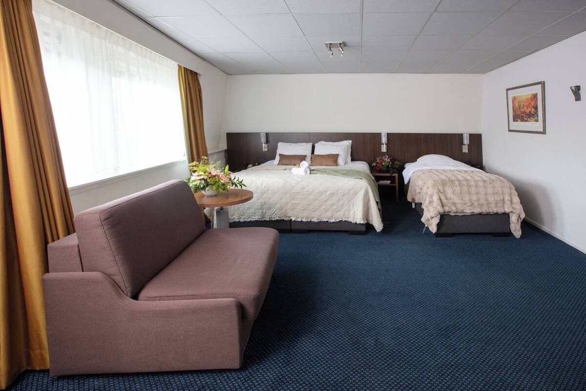 A hotel room with two beds, a sofa, a small table with flowers, and a painting on the wall.