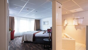 A hotel room with a large bed, a small round table, a desk, a chair, a window with curtains, a bathroom with a bathtub, and towels on a rack. - Petit Nord (Hoorn)