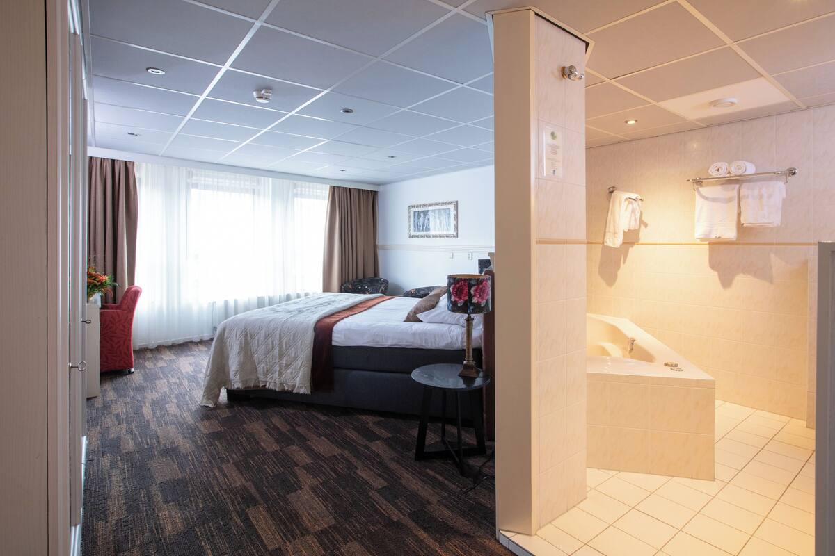 A hotel room with a large bed, a small round table, a desk, a chair, a window with curtains, a bathroom with a bathtub, and towels on a rack.