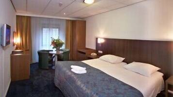 Habitación de hotel con una cama grande, una mesita de noche, un televisor, un cesto de basura y una ventana con cortinas.