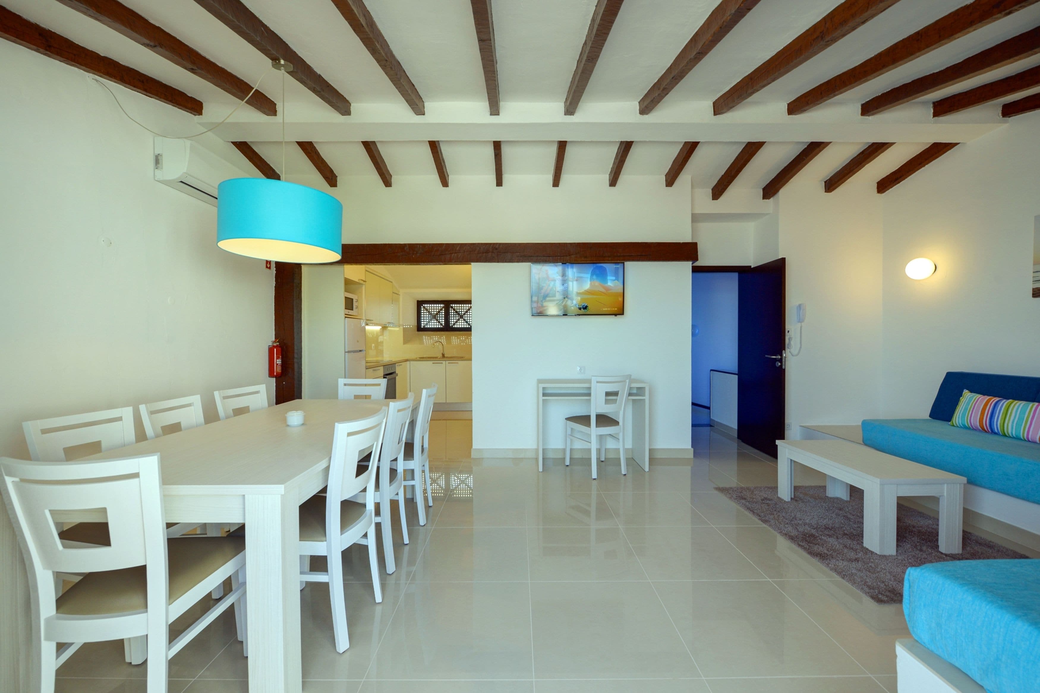 A modern living room with a dining area, featuring a blue sofa, white furniture, and a kitchen in the background.