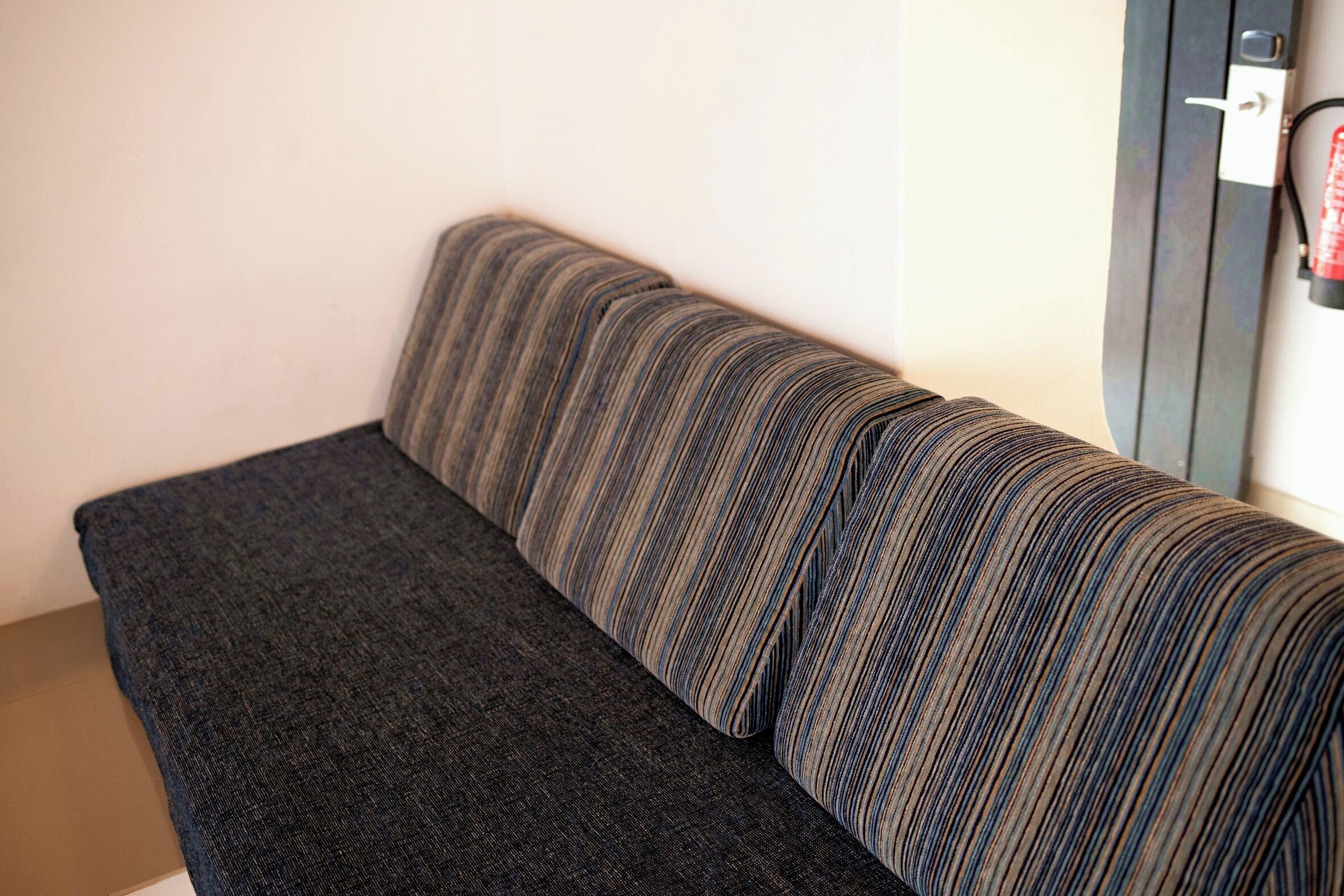 A sofa with a striped pattern in a room with a plain wall and a door with a fire extinguisher mounted on it.