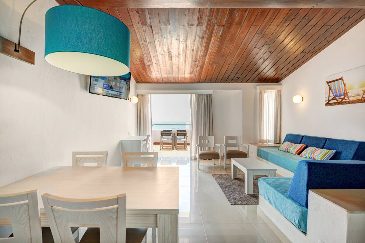 Un salon moderne avec un plafond en bois, un canapé bleu, une table à manger avec des chaises et une vue sur l’extérieur.