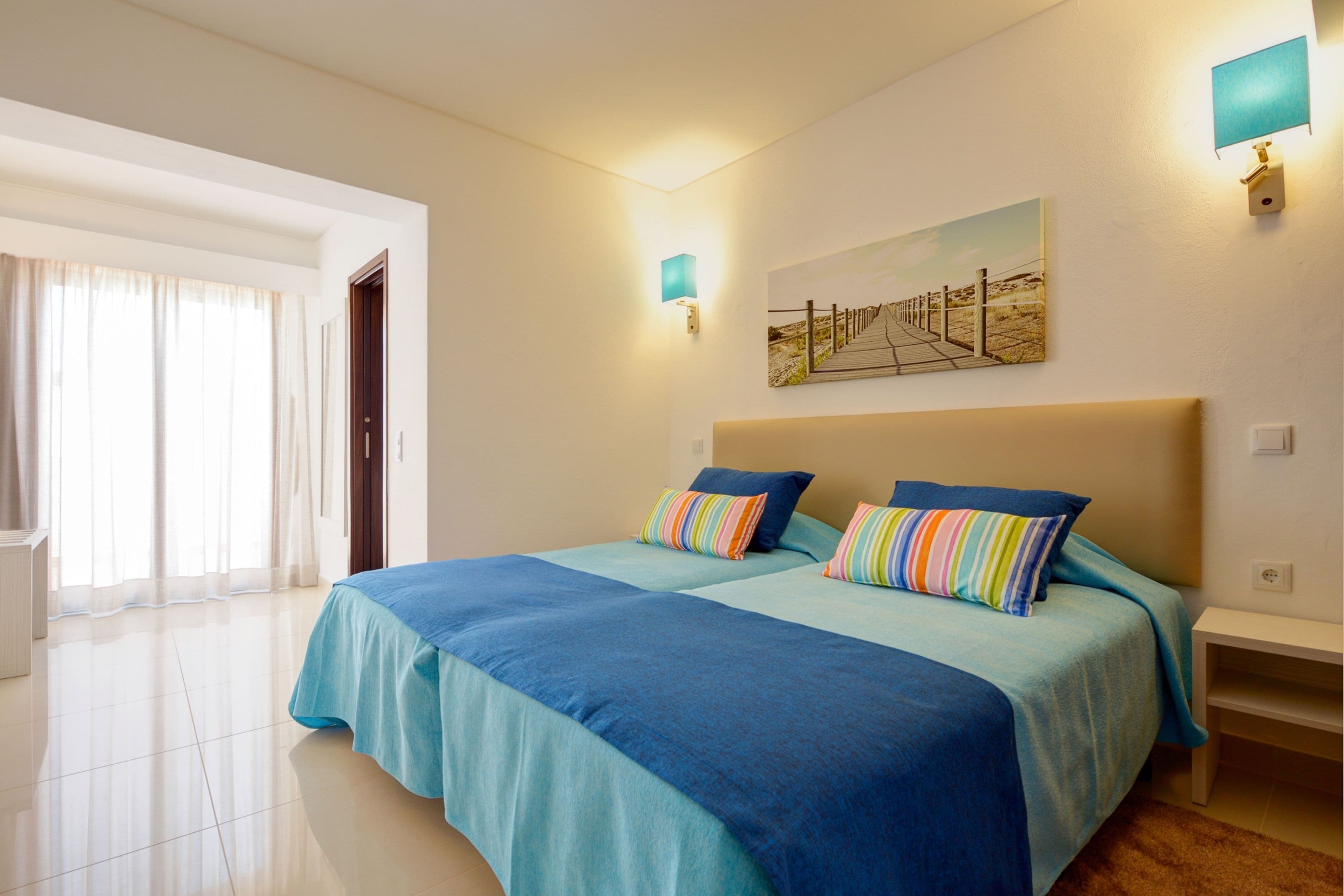 A hotel room with two beds, a large window with curtains, a painting on the wall, and a nightstand.