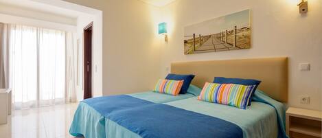A hotel room with two beds, a large window with curtains, a painting on the wall, and a nightstand.
