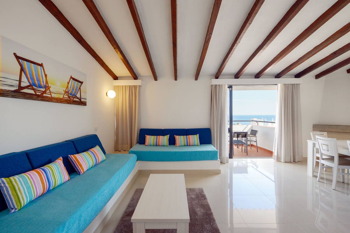 Un salon moderne avec un canapé bleu, une table basse blanche et une vue sur la mer depuis un balcon.