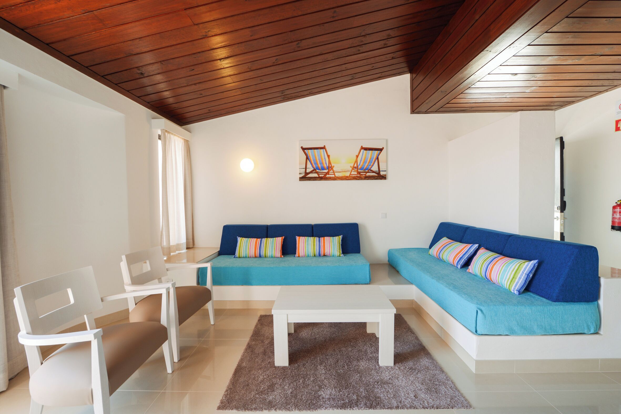 Una sala de estar con techo de madera, un sofá azul, una mesa de centro blanca y dos sillas blancas.