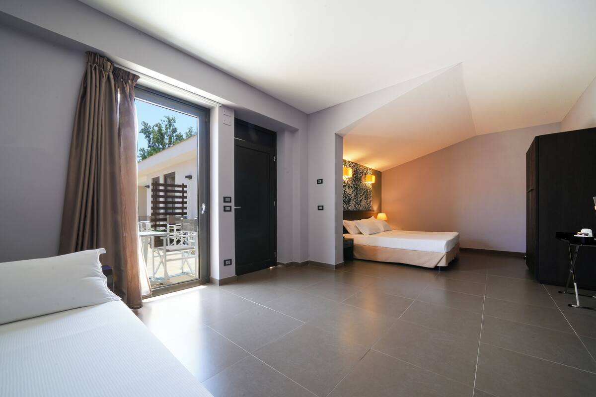 A modern hotel room with a large bed, a smaller bed, and a balcony with a dining area.