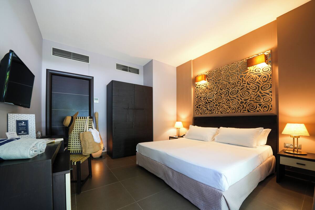 A hotel room with a large bed, two bedside tables with lamps, a wardrobe, a desk with a chair, and a television on the wall.