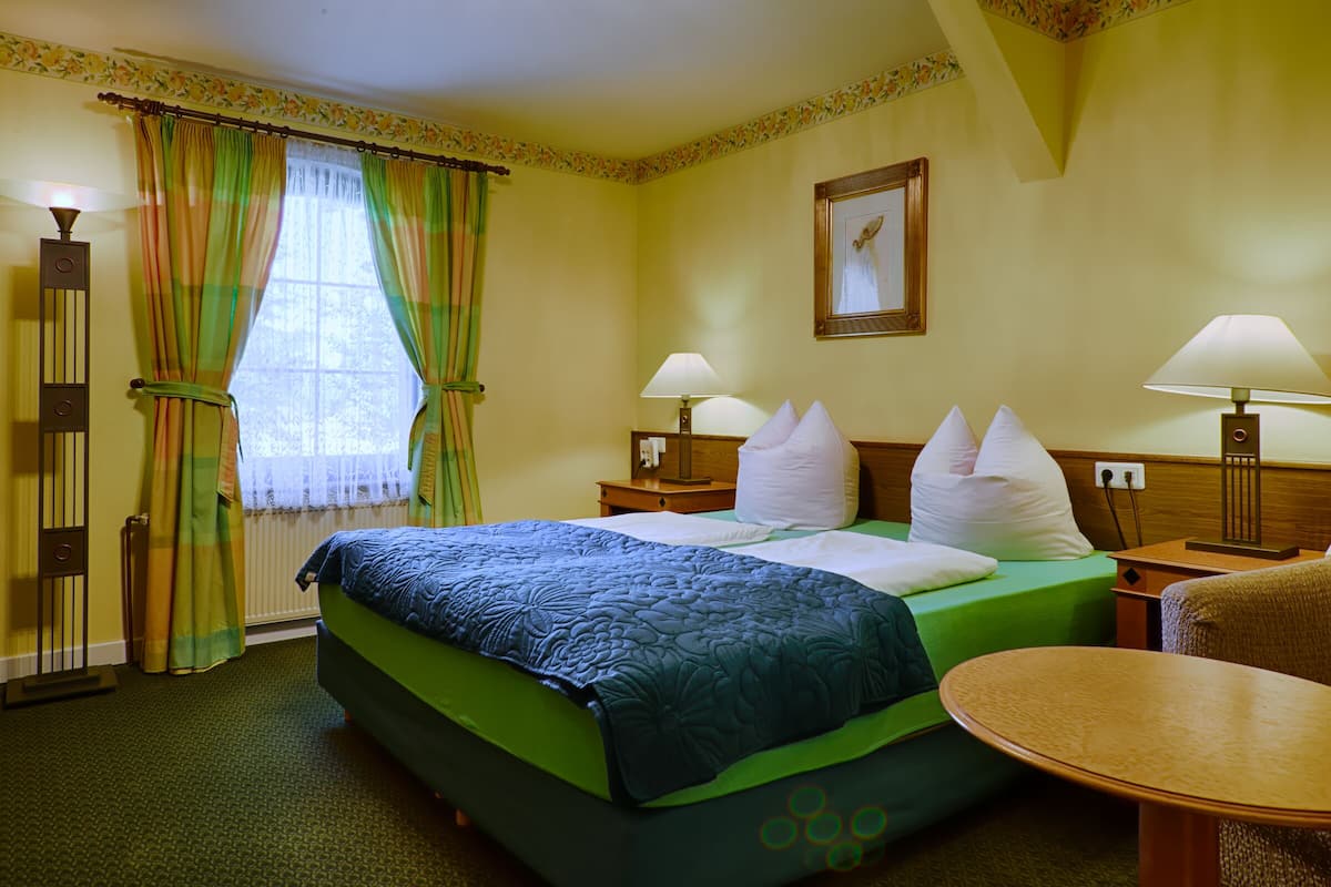 Ein Hotelzimmer mit einem Bett, zwei Nachttischen mit Lampen, einem Sessel, einem runden Tisch und einem Fenster mit Vorhängen.