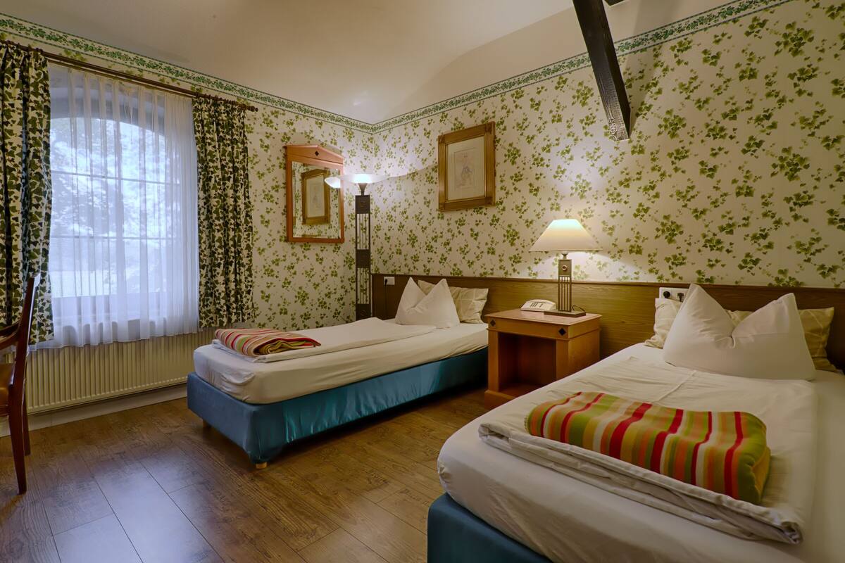 Ein Hotelzimmer mit zwei Betten, Holzboden, Heizkörper, einem Fenster mit durchsichtigen Vorhängen, einem Nachttisch mit Lampe und floralem Wandpapier.