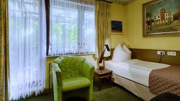 Una habitación de hotel con una cama, un sillón verde, una mesita de noche con una lámpara y un cuadro en la pared.