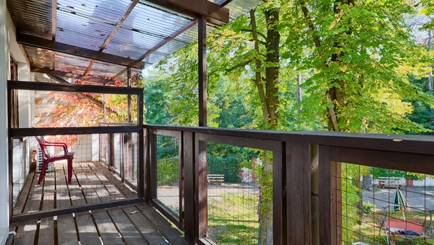Une terrasse en bois avec une rambarde en métal, une chaise rouge et une vue sur un parc verdoyant, avec des arbres et un banc.