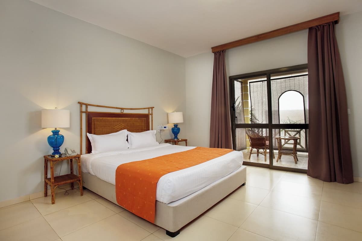 A hotel room with a large bed, two bedside tables with lamps, a view of the outdoors through a glass door, and a balcony with wicker furniture.