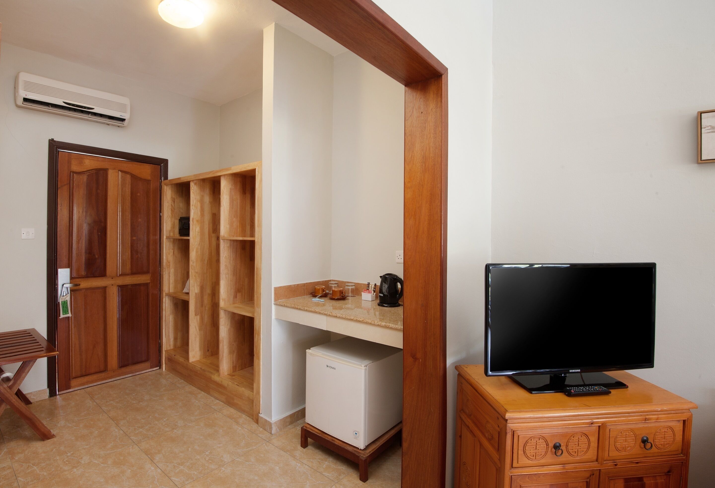 Una habitación pequeña con un armario de madera, un refrigerador compacto, un escritorio de madera con un televisor y una estantería con diversos objetos.