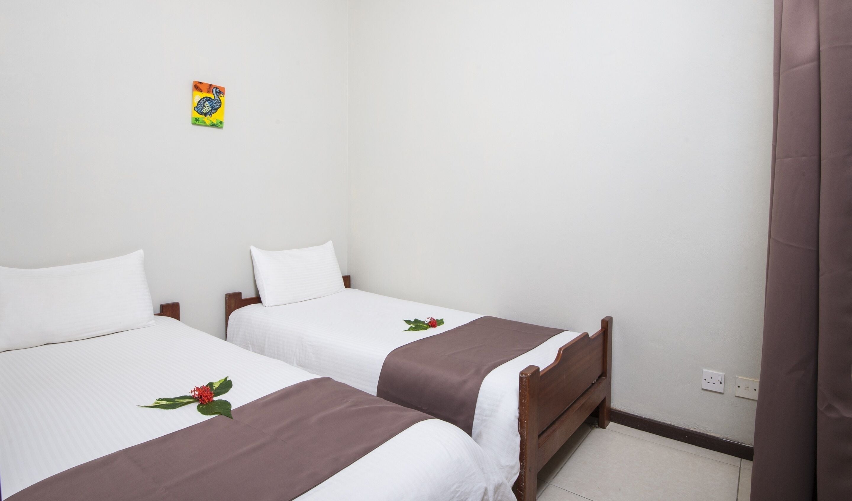 Habitación doble con dos camas, cada una con sábanas blancas y colchas marrones, un pequeño adorno en la pared y una cortina a la derecha.
