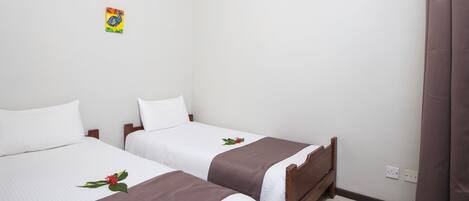 Habitación doble con dos camas, cada una con sábanas blancas y colchas marrones, un pequeño adorno en la pared y una cortina a la derecha.