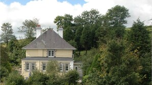 A two-story house with a gabled roof, surrounded by dense greenery and trees. - The Oratory (Yelverton)
