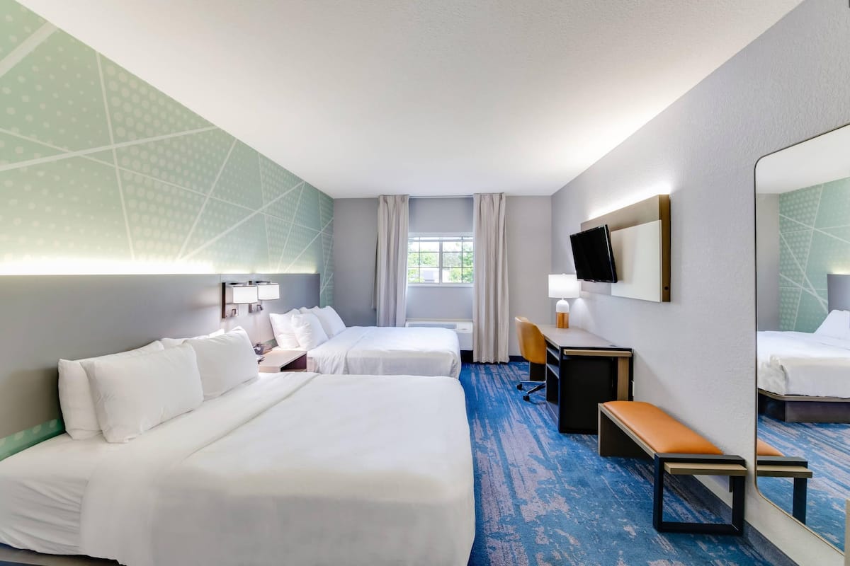 Habitación de hotel con dos camas, un escritorio, una silla, un banco, un televisor y un espejo grande.
