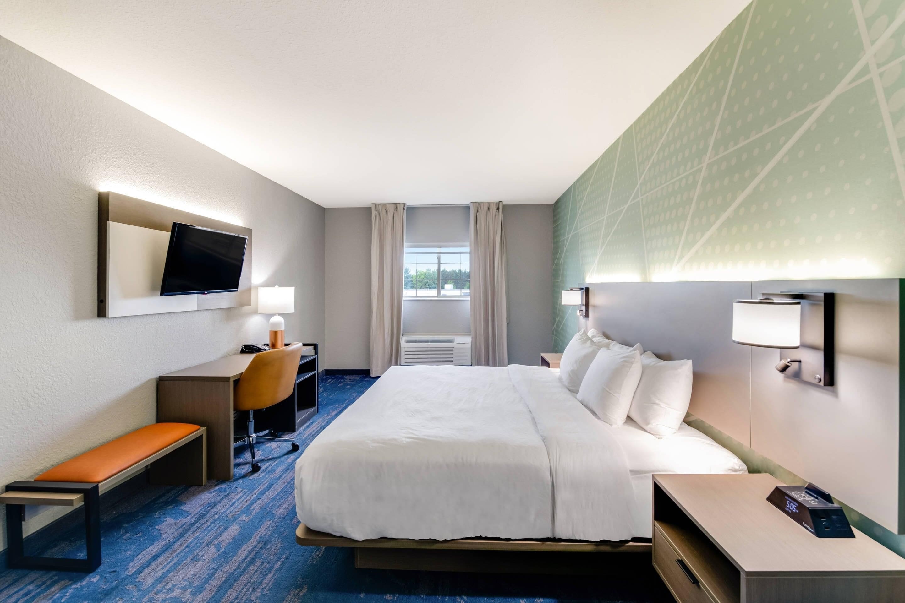 A hotel room with a large bed, a desk with a chair, a bench, a TV mounted on the wall, and a window with curtains.