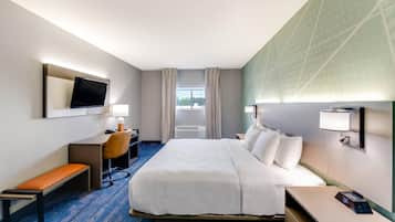 A hotel room with a large bed, a desk with a chair, a bench, a TV mounted on the wall, and a window with curtains.