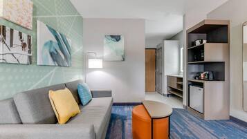 A modern hotel room with a grey sofa, a round orange ottoman, a blue and orange patterned carpet, a wall with abstract art, and a kitchenette area.