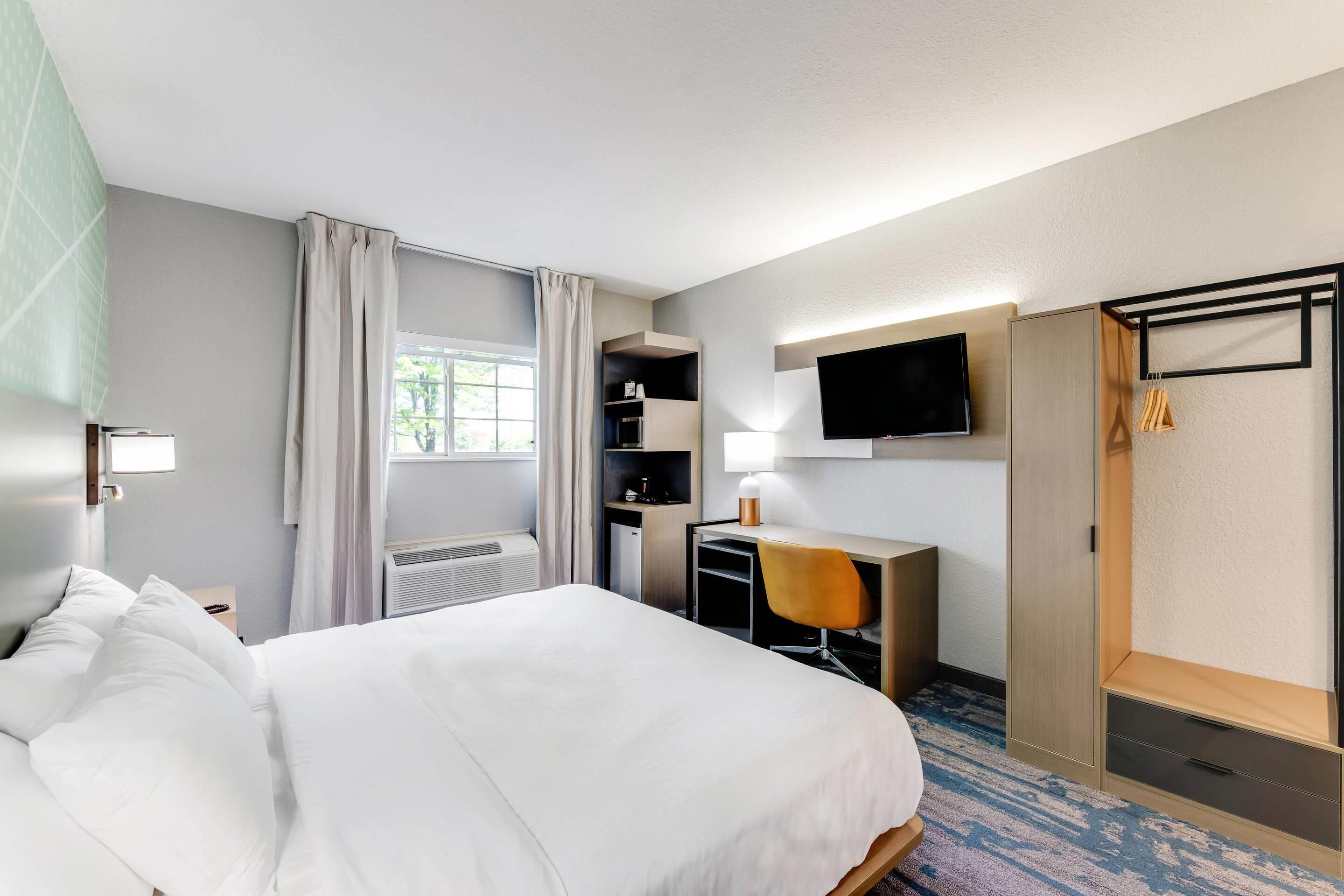 A hotel room with a bed, desk, chair, TV, wardrobe, and a window with curtains.