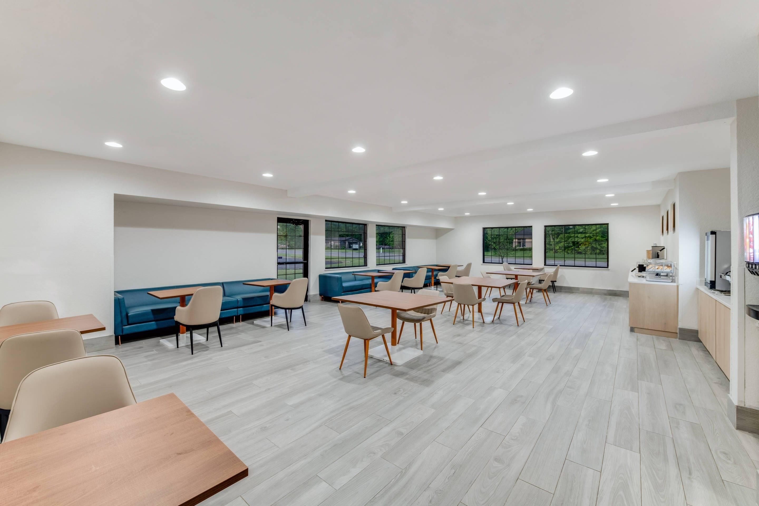 A modern dining area with wooden tables and chairs, a blue sofa, and large windows providing natural light.