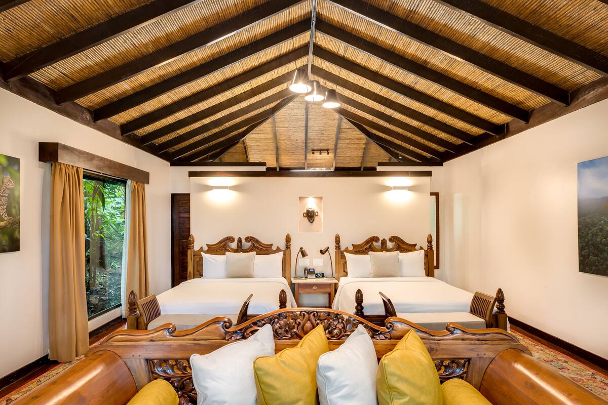 A hotel room with a high ceiling, two beds, a wooden headboard, a nightstand, and a view of greenery through a window.