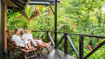 A wooden deck with two people seated on a bench, surrounded by lush greenery and a wooden railing.